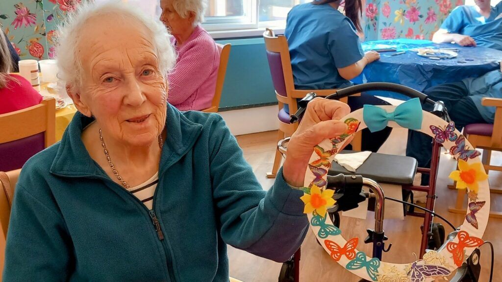 During Craft Afternoon, Celia sits at a table holding a fabulous craft—a decorated wreath with colourful butterflies and flowers—in a communal room with others in the background.