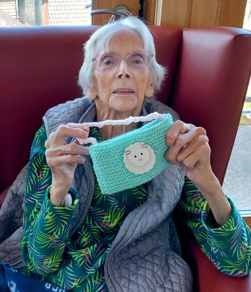 An elderly woman sitting in a red armchair holds up a small blue knitted pouch with a smiling face patch, proudly displaying her handiwork from the Knit and Natter knitting group.