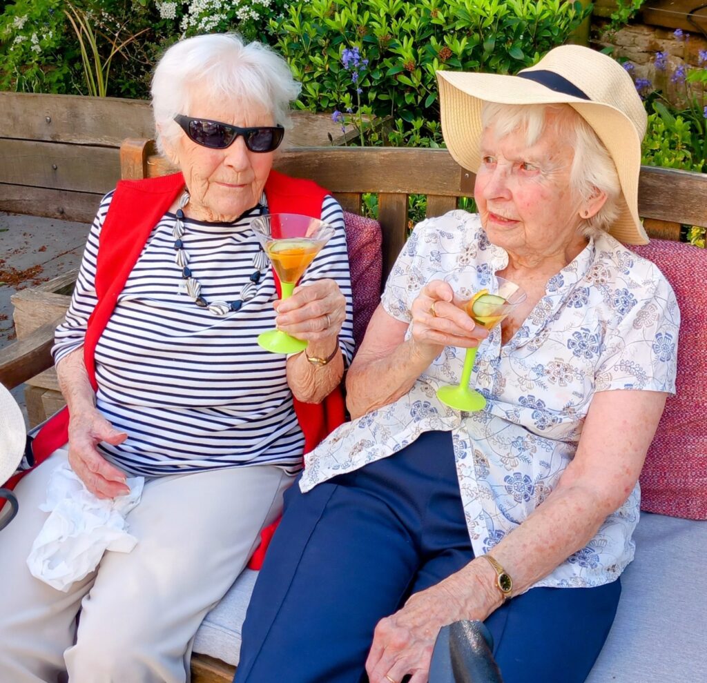 Two elderly women enjoy good company on an outdoor bench, holding Pimm’s in stemmed glasses. One wears sunglasses and a red waistcoat, the other a sunhat and floral blouse.