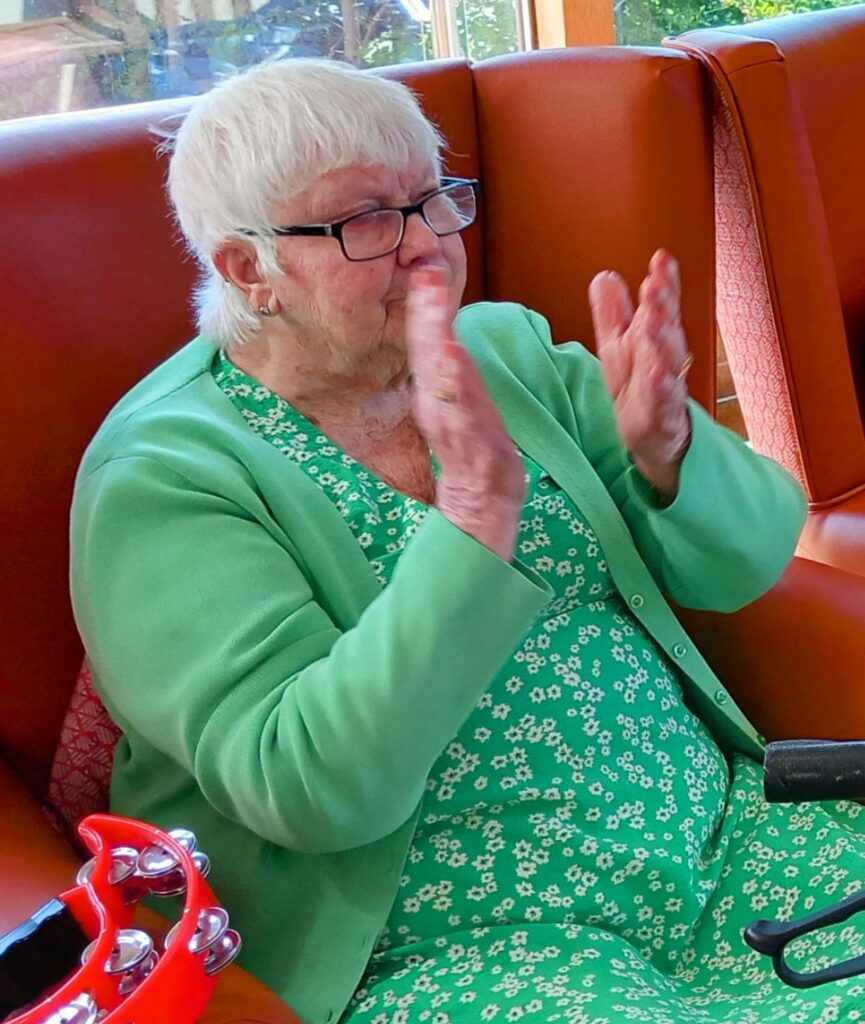 Elderly woman with short white hair and glasses, dressed in a green dress and cardigan, claps her hands while seated on a red chair; a red tambourine rests on her lap during the VE Day 80th Anniversary celebration.
