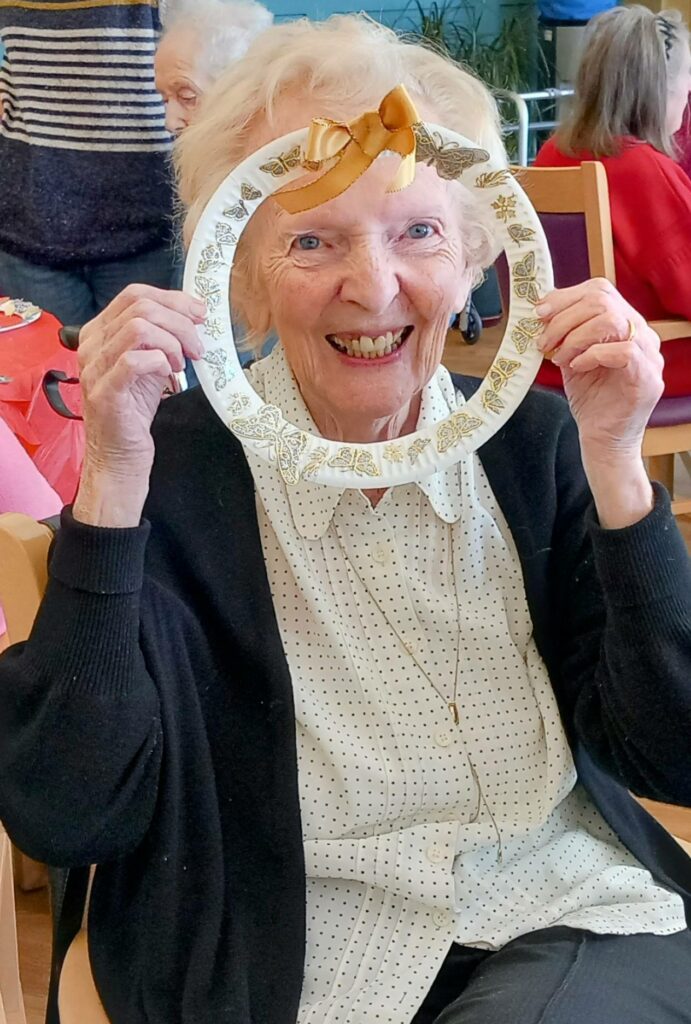 Elderly woman Celia smiling and holding a decorative paper wreath in front of her face while seated at a table during a craft workshop in a communal setting.