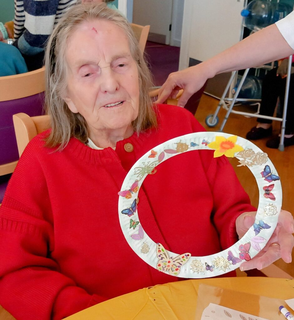 Celia, an elderly woman in a red jumper, enjoys Craft Afternoon as she holds a decorated circular craft featuring butterfly and flower designs, seated at a table indoors.