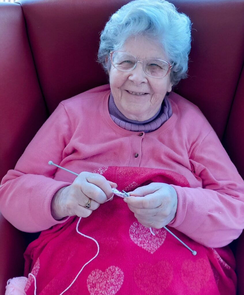 An elderly woman with grey hair and glasses sits on a red chair, smiling as she knits with pink wool—perhaps preparing for the upcoming April Knit, a cheerful knitting event she greatly enjoys.