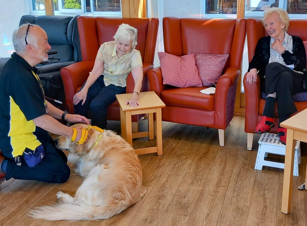 During a Tim Visit, a man kneels beside a golden retriever wearing a yellow hat, while two elderly women sit in armchairs, smiling and watching the dog in a brightly lit room.