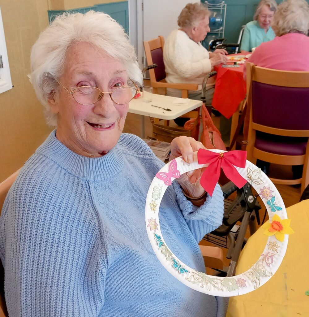 During Craft Afternoon, Celia, an elderly woman, sits at a table holding a decorated paper wreath with a red bow whilst others work on crafts in the background.