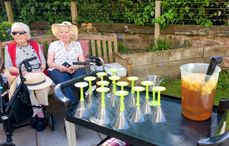 Two elderly women sit in good company on a garden bench beside a table with plastic glasses and a jug of Pimm’s, enjoying the outdoors on a sunny day.