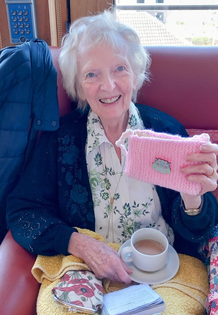 An elderly woman sits on a red chair at a Knit and Natter, holding a pink knitted pouch and a cup of tea, with a notepad on her lap and a jacket draped over the chair.