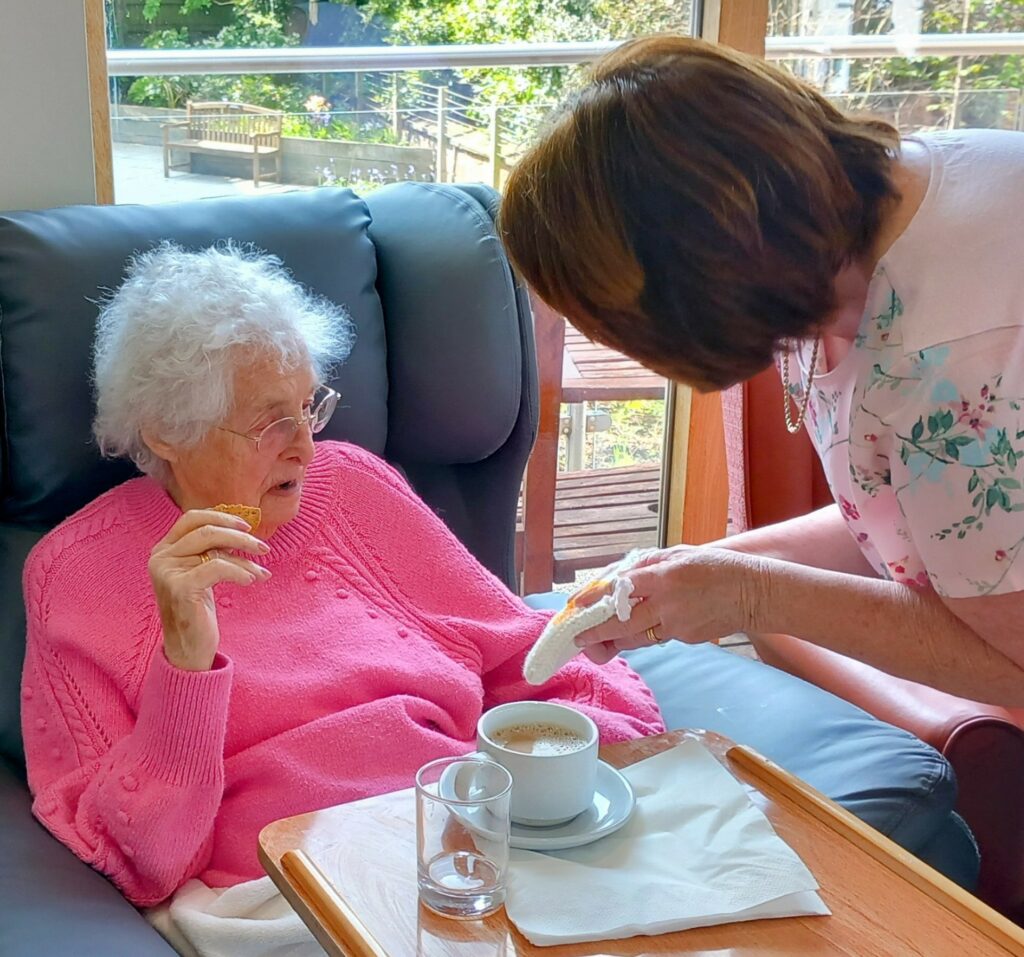 An elderly woman sits in an armchair holding a biscuit while another offers her a serviette at a Knit and Natter knitting event, with coffee and water served on a nearby tray.