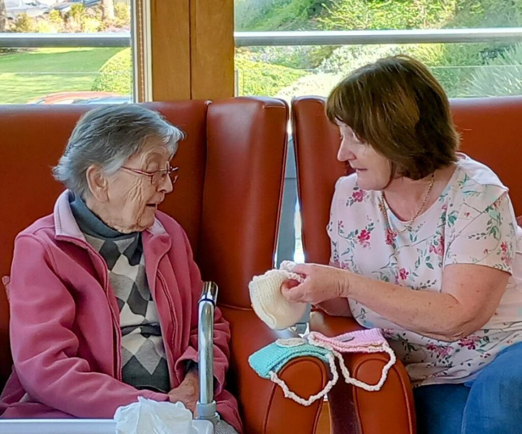 An elderly woman and a middle-aged woman are sitting on a sofa; the middle-aged woman is showing off April knitting projects from their Knit and Natter group.