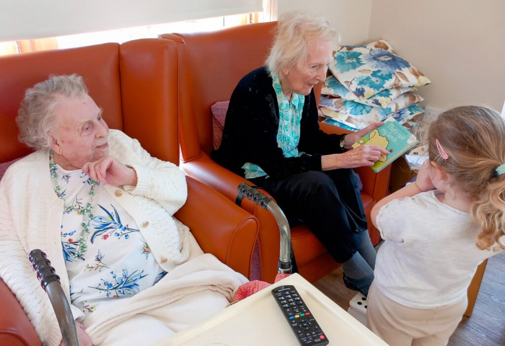 Two elderly women sit in armchairs; one holds a children’s book, sharing laughter with a young girl nearby. A stack of cushions and a TV remote add comfort to this cosy moment with little ones.