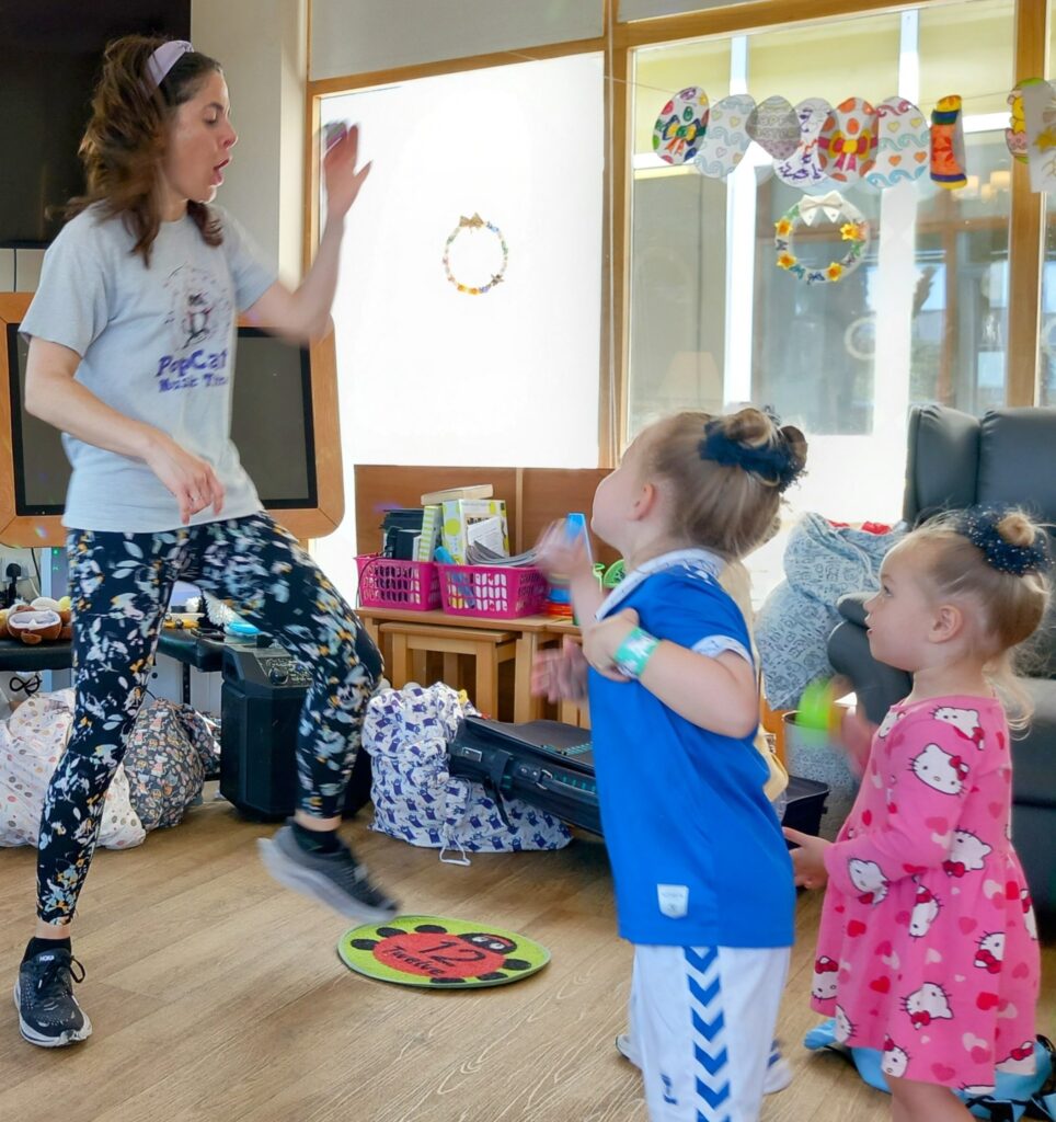 An adult leads some little ones in an indoor activity; laughter fills the room as she raises her hand and leg, while the children watch and copy her movements.