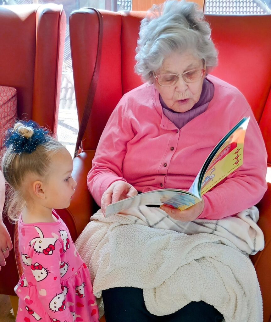 An elderly woman wearing glasses reads lyrics from a book to a young girl in a pink Hello Kitty dress. Laughter fills the air as they sit together on a red chair with a light blanket.