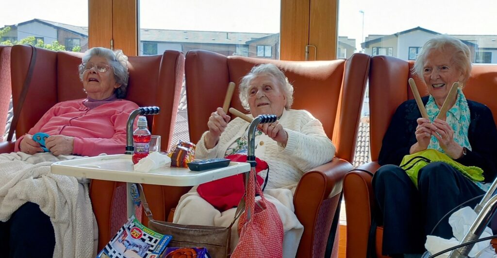 Three elderly women sit in armchairs by a window, each holding claves and smiling with laughter. Walking frames and personal belongings are close by.