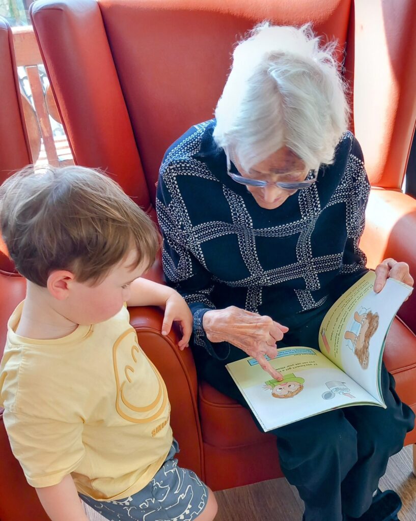 An elderly woman sits in an armchair, reading a picture book to a young boy. Laughter fills the room as she points to an illustration, delighting the little ones with each page.