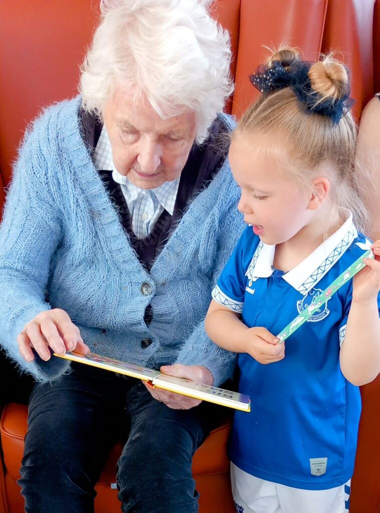 An elderly woman and a young girl share laughter on a red seat while looking at a book. The girl, in a blue sports kit, holds a pencil as they enjoy special moments together.