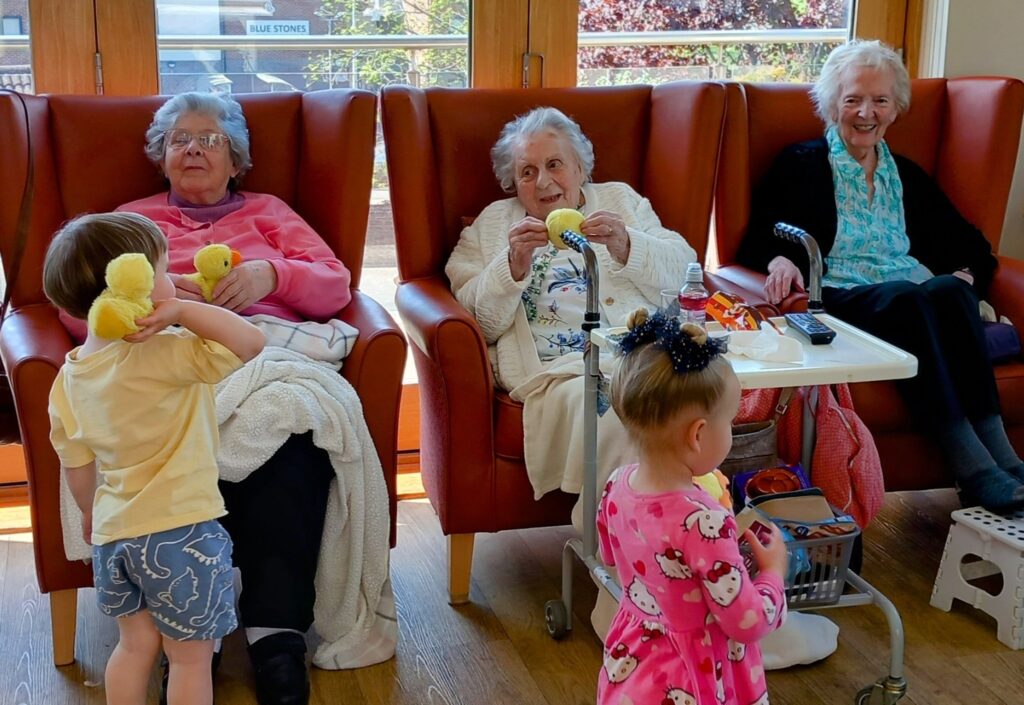 Three elderly women sit in armchairs, while two little ones stand in front; sunlight streams through large windows behind the group, filling the room with warmth and laughter.