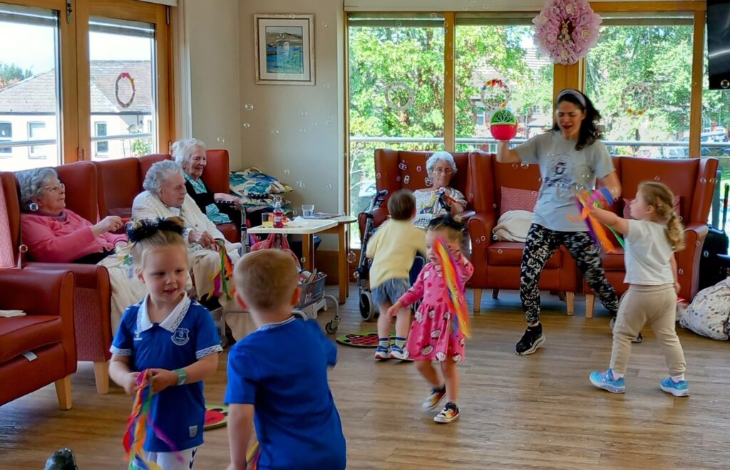 Laughter fills the brightly lit community room as young children and elderly women play with streamers and bubbles near large windows.