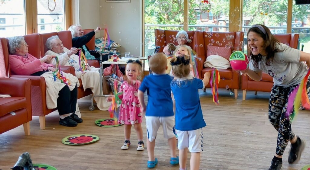 A group of elderly people sit in armchairs, laughter filling the bright communal room as they watch three little ones and a woman with ribbons taking part in an activity together.