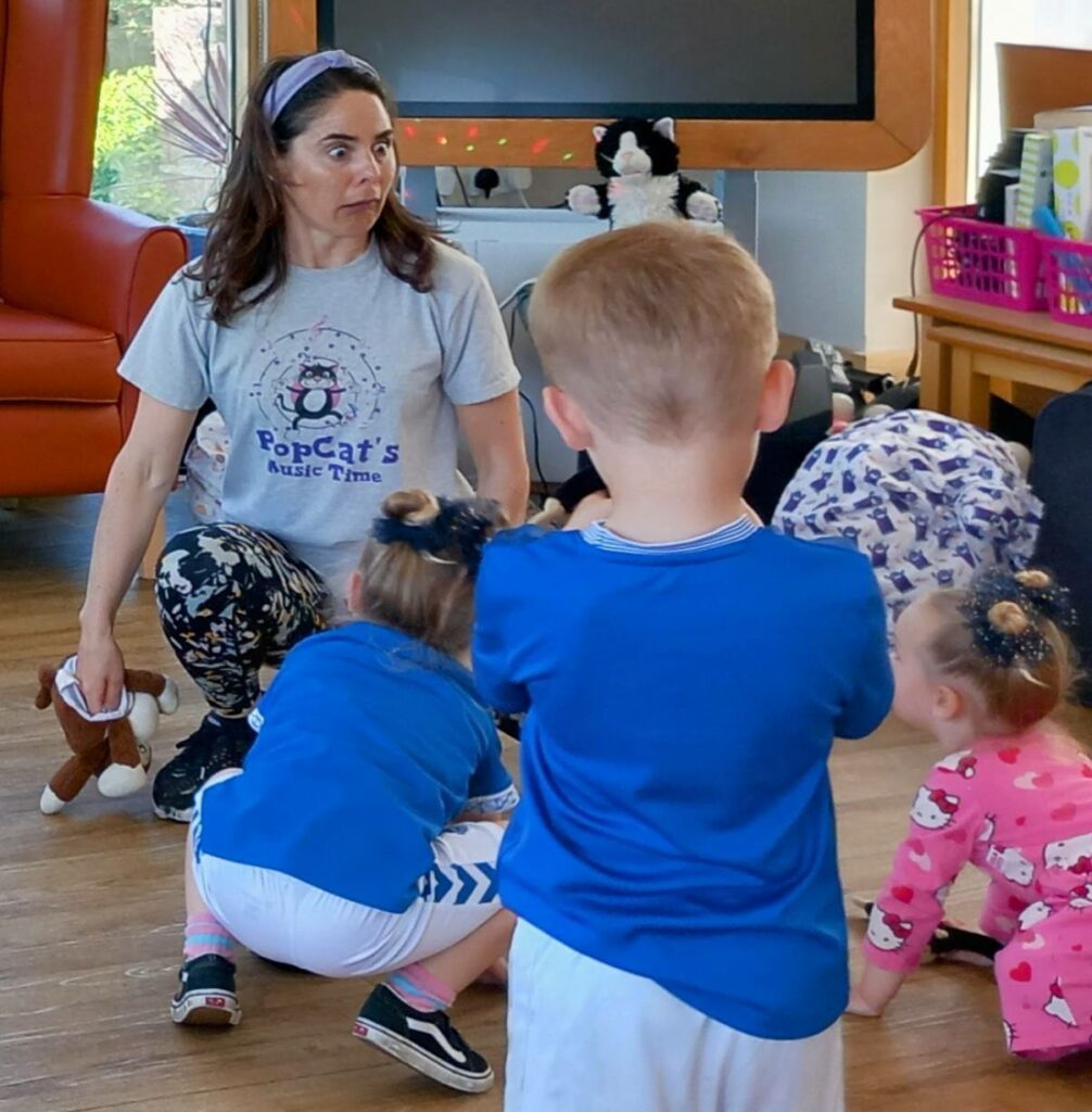 A woman wearing a PopCats Music Time shirt leads young children in an indoor activity on a wooden floor. The children, dressed in colourful clothes, share smiles and laughter throughout the enjoyable session.