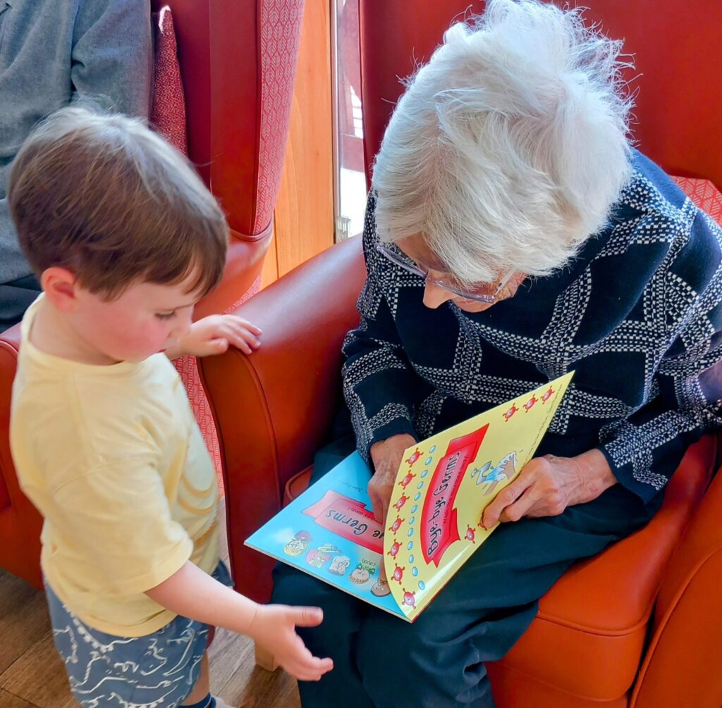 An elderly woman sitting in a red armchair reads a colourful children’s book to a young boy standing beside her, filling the room with laughter and delight for the little ones.