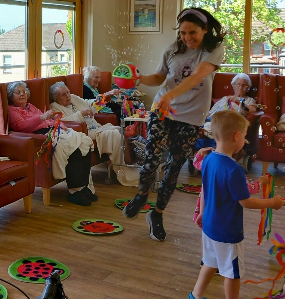 A woman leaps into the air holding a ball while older adults sit and watch; laughter fills the brightly lit room, decorated with ladybird mats, as a young boy stands nearby.