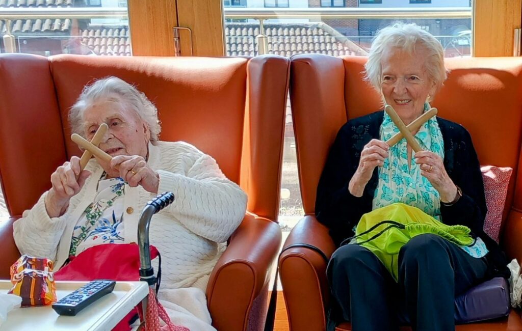 Two elderly women sitting in armchairs, each holding two wooden sticks crossed over, smiling at the camera, their laughter echoing like lines from treasured memories.