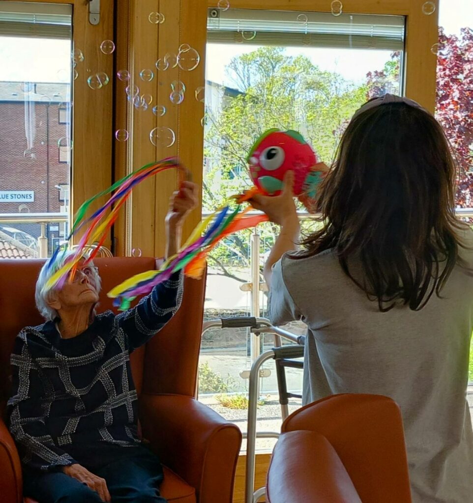 An older woman seated in an armchair reaches towards colourful streamers on a toy fish held by another person; bubbles float around them indoors, filling the moment with laughter.