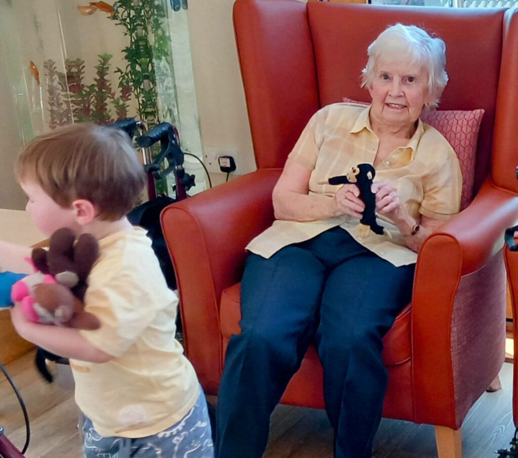 An elderly woman sits in a red armchair, holding a small soft toy, while laughter fills the room as a young child—one of the little ones—walks past, arms full of cuddly toys.