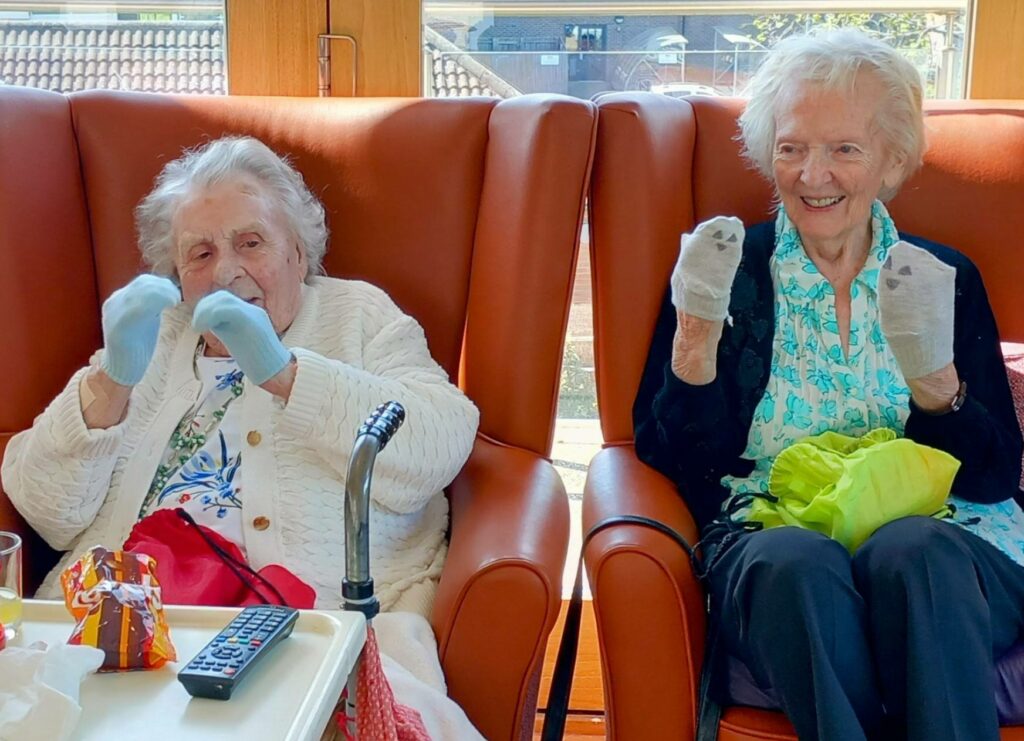 Two elderly women sit side by side in armchairs, smiling and holding up their hands with white socks as mittens, sharing laughter. A walking stick and remote control rest on the table in front of them.
