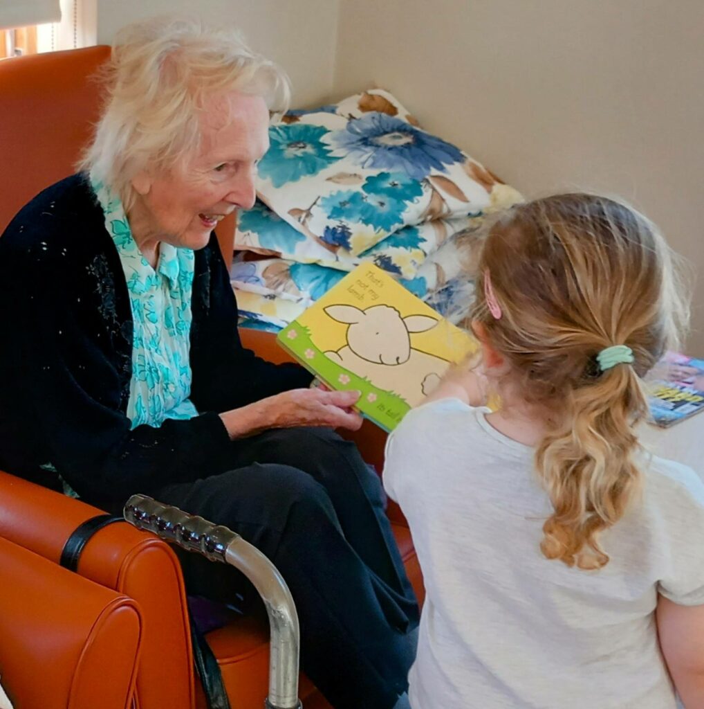 An elderly woman sits in an orange chair, smiling at a young girl from the Little Ones who is showing her a children’s book with a sheep on the cover, filling the room with laughter.