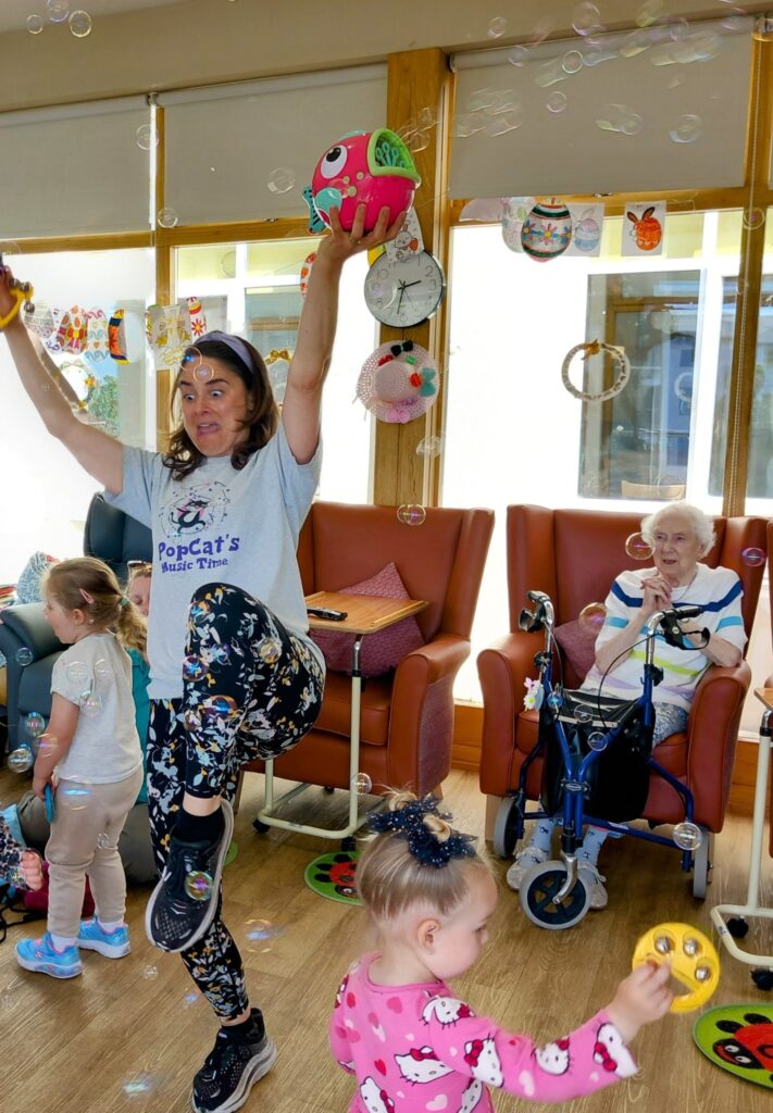 A woman leads a joyful music and movement session for young children and elderly residents in a care home, with laughter filling the air, bubbles floating about, and colourful props in use.
