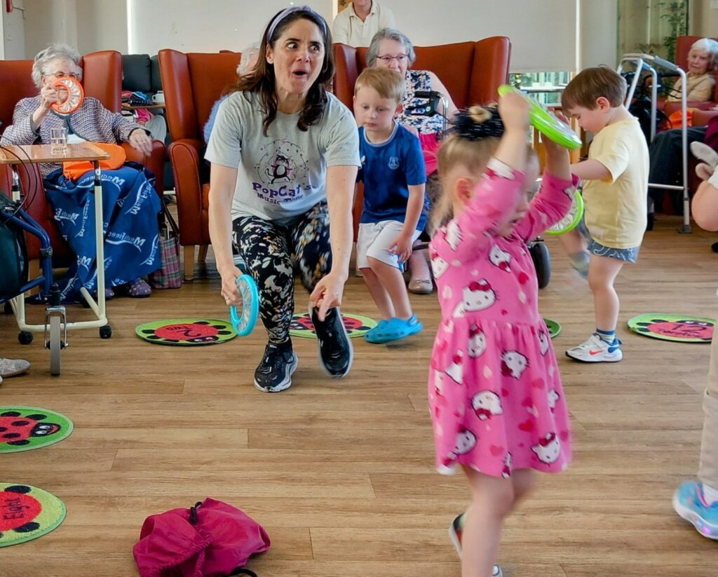 A woman leads Little Ones in a lively activity with older adults in a brightly lit room. Children, full of laughter, wave colourful rings energetically as the seniors watch from their armchairs.