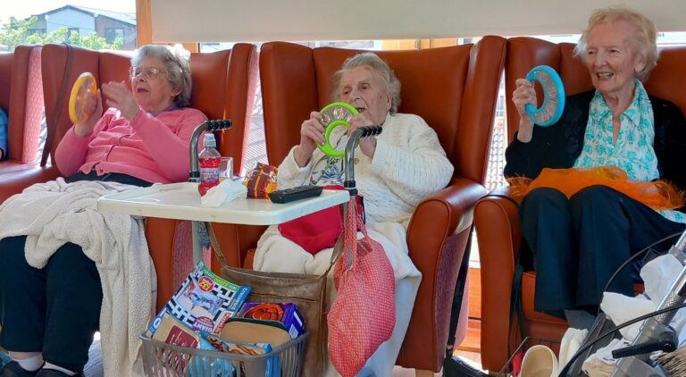Three elderly women sit in armchairs, laughter filling the well-lit room as they hold coloured plastic rings, with walking frames and personal belongings nearby.