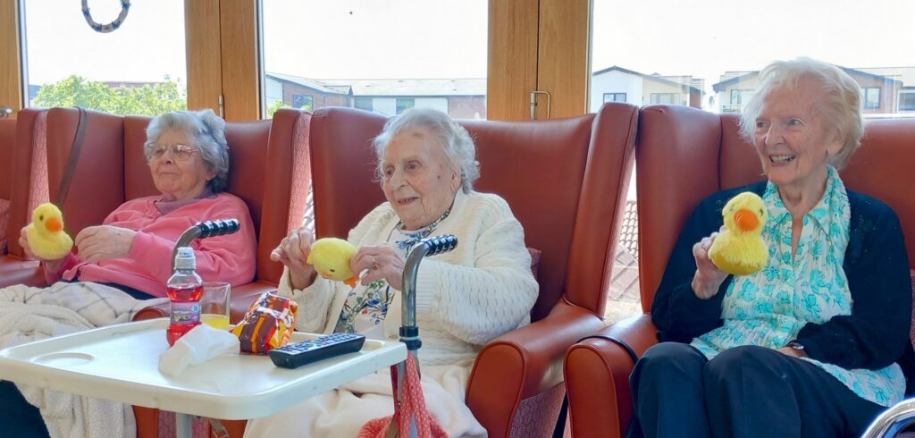 Three elderly women sit in armchairs by large windows, each holding a yellow rubber duck, their laughter filling the room as snacks and a remote control rest on the tray in front of them.