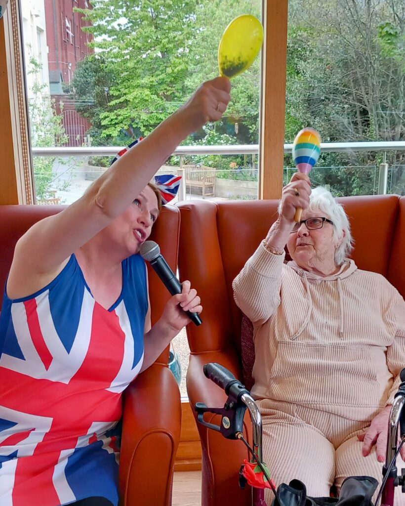 Two women seated in armchairs shake maracas; one holds a microphone and wears a Union Jack dress, while the other, wearing glasses and beige clothing, sits with a walking frame beside her.