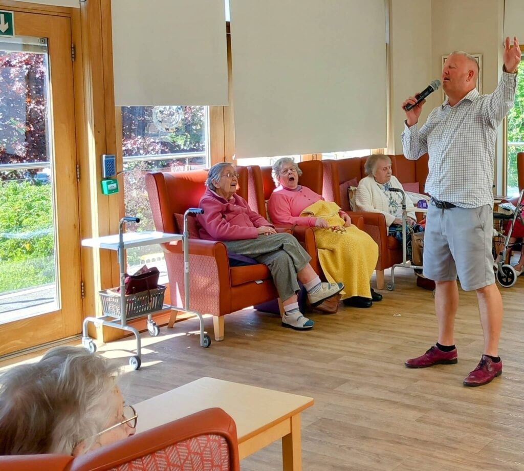 A man is singing into a microphone while four elderly women sit and watch him in a brightly lit room with large windows.