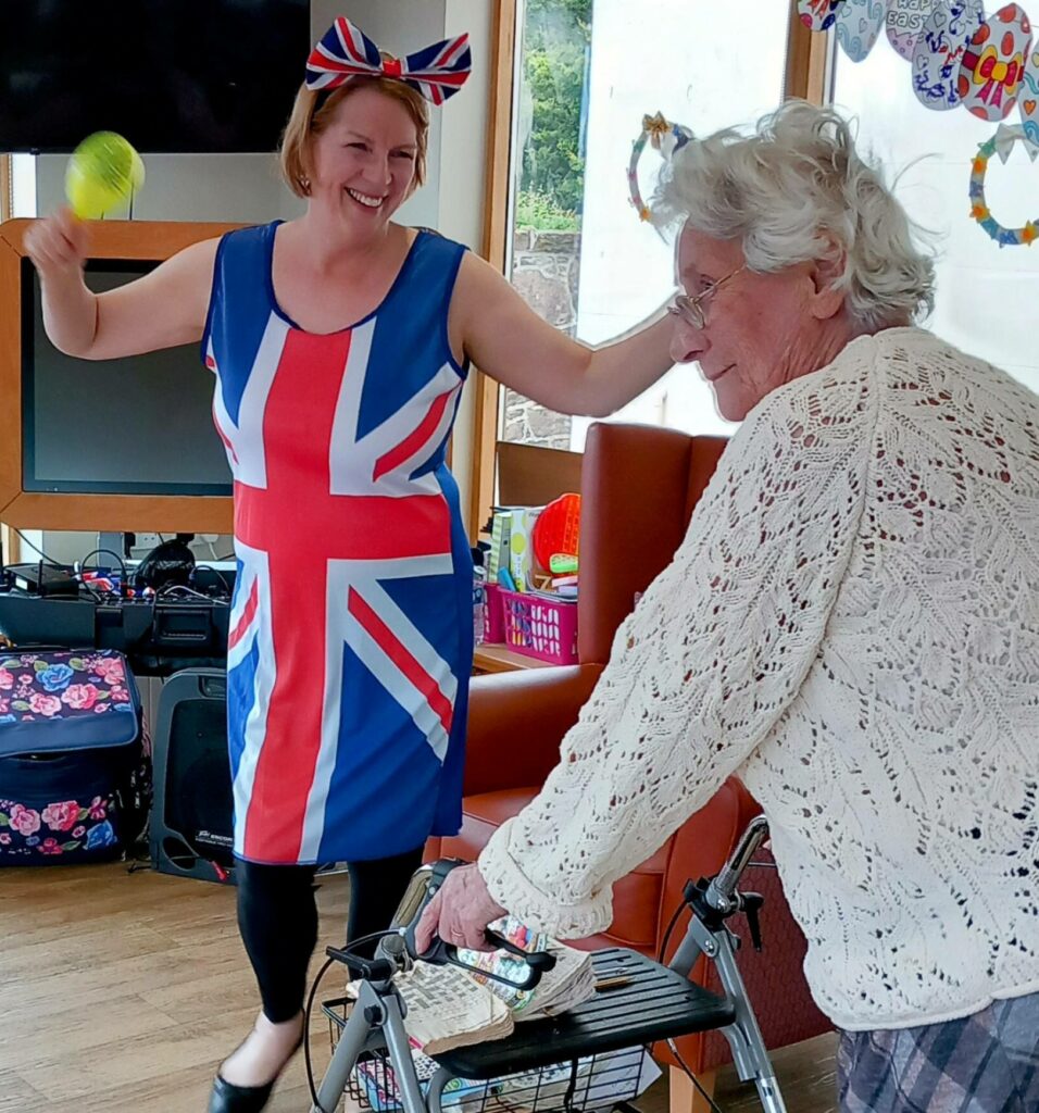 A woman in a Union Jack dress and bow dances with a maraca beside an elderly woman using a walking frame in a brightly lit room.