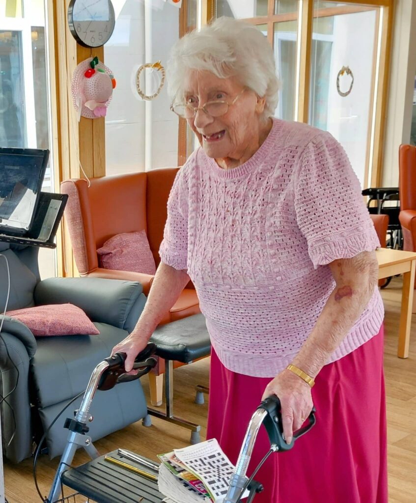 Elderly woman with white hair and glasses uses a walking frame indoors, wearing a pink knitted top and skirt. She is smiling, perhaps singing along to a tune, with chairs and a clock in the background.