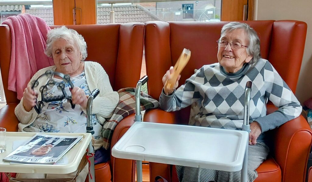 Two elderly women sit in chairs, smiling and playing musical instruments; one holds a tambourine, the other a shaker. Both have walking frames and trays in front of them.