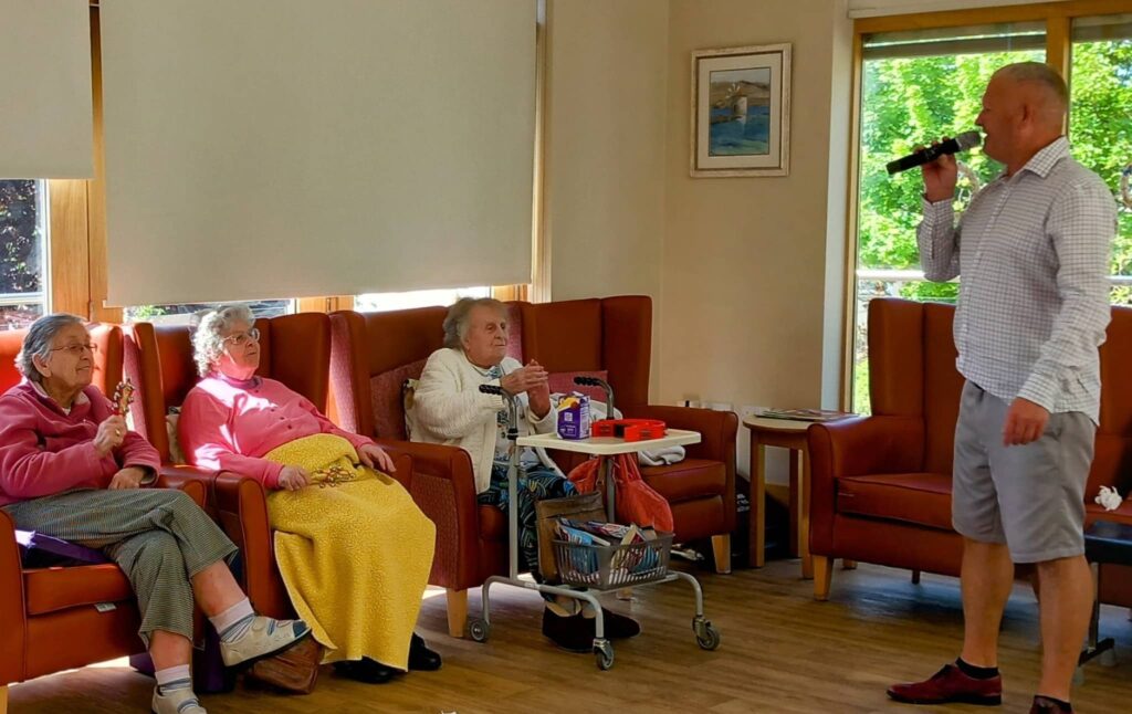A man is singing into a microphone in a brightly lit room while three elderly women sit in armchairs, listening.