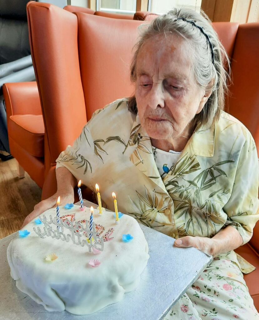An elderly woman sits in front of a birthday cake with five lit candles, preparing to blow them out. Happy Birthday, Joan!.