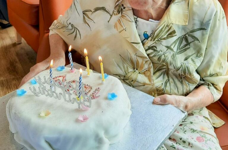 An elderly woman sits in front of a birthday cake with five lit candles, preparing to blow them out. Happy Birthday, Joan!.