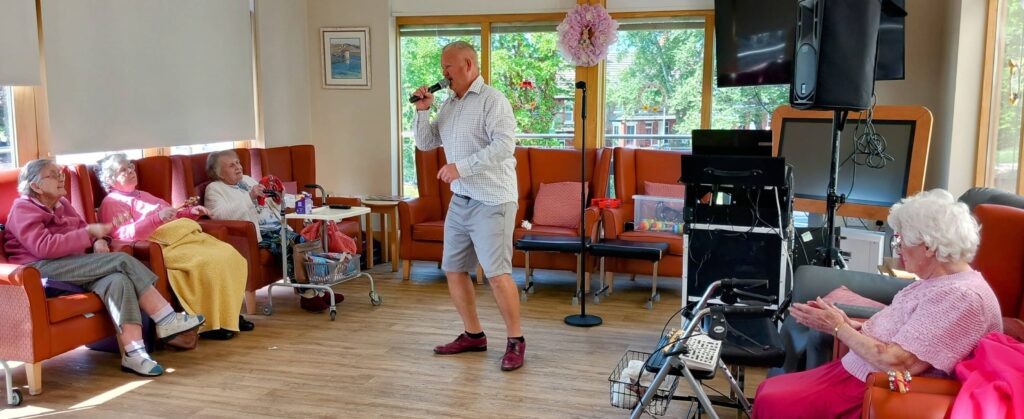 A man is swinging and singing into a microphone in a brightly lit room while several elderly people seated in chairs watch and clap.