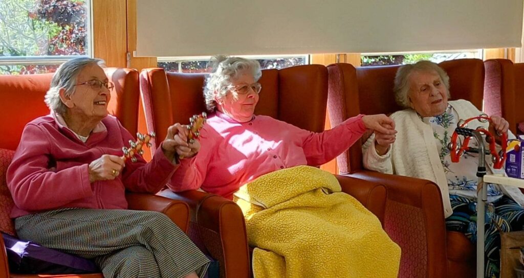 Three elderly women sit on red chairs, holding hands and musical instruments, singing together as sunlight streams through the windows behind them.