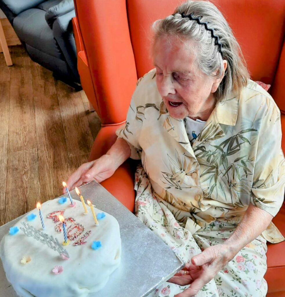 An elderly woman sits in an orange armchair, gazing at a birthday cake with lit candles and 94 written on it. Happy Birthday, Joan!.