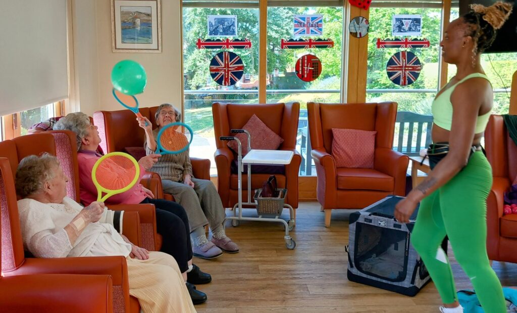 A group of elderly women enjoy a lively Active Afternoons session as Carla, dressed in green exercise kit, leads them in hitting balloons with bats in a bright room decorated with British flags.