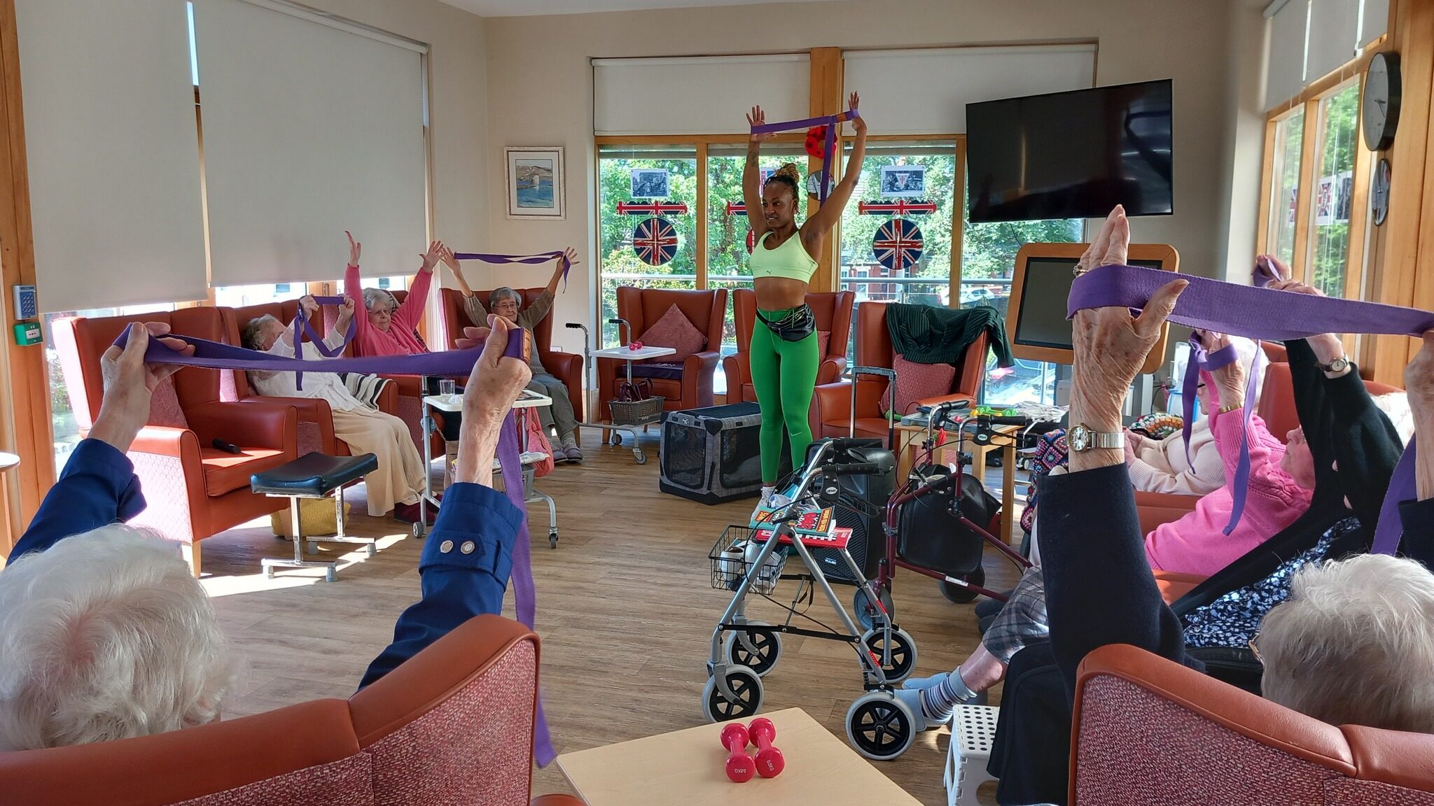 Active Afternoons: A group of elderly people in armchairs follow an exercise instructor, stretching with resistance bands and sharing smiles with Carla in a bright activity room.