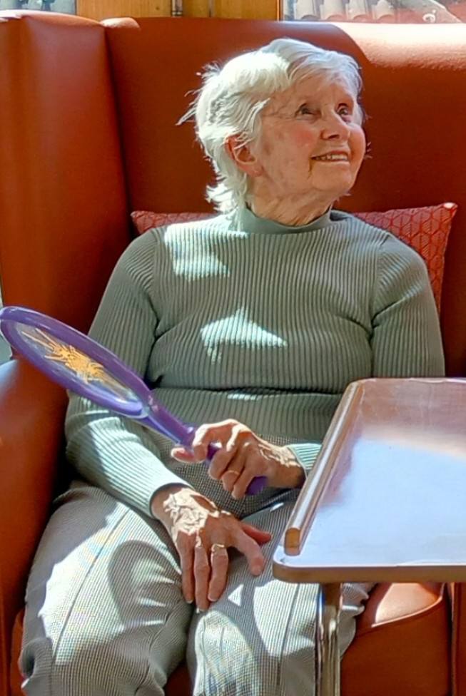 An elderly woman with short grey hair sits in an orange chair, holding a purple handheld mirror and looking up with a slight smile during Active Afternoons.