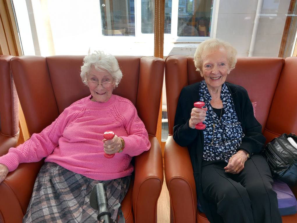 Two elderly women sit in armchairs, each holding a small pink dumbbell, smiling at the camera during Carla’s Active Afternoons in a bright indoor setting.