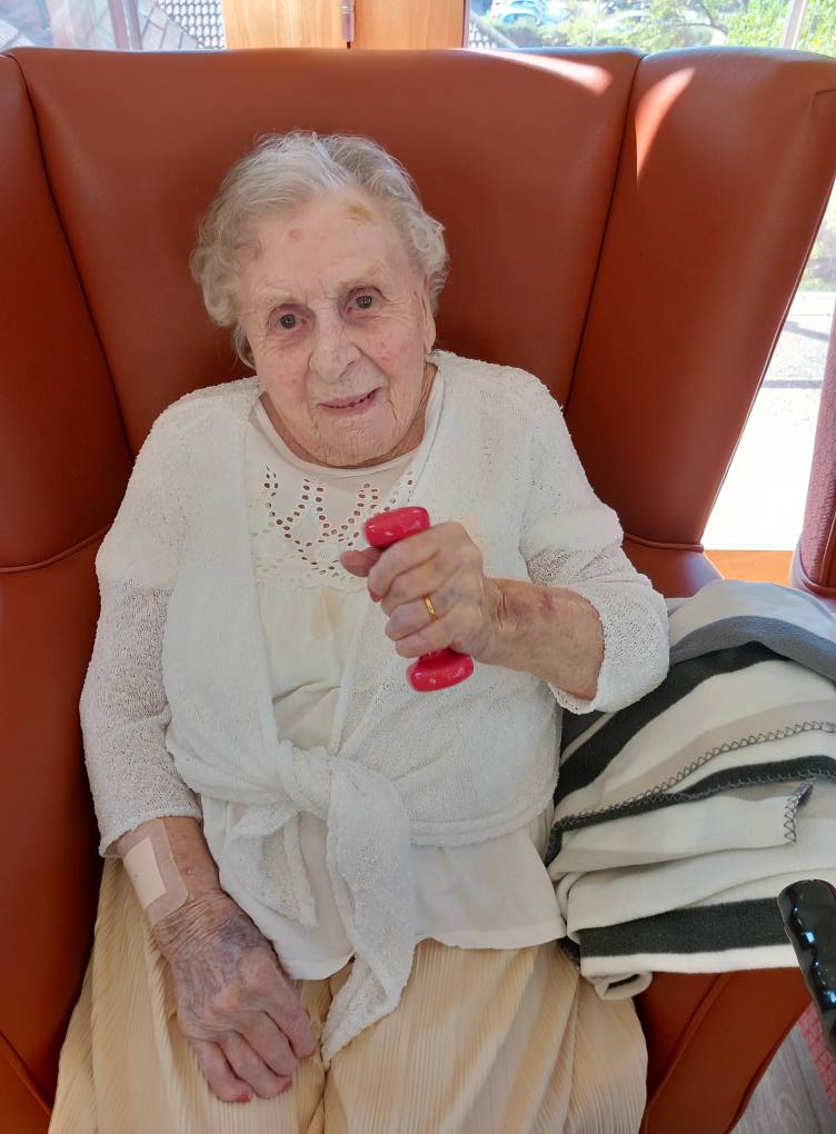 An elderly woman sits in a cushioned chair during Active Afternoons, holding a small red dumb-bell in her right hand. She is dressed in white, with a blanket and walking stick beside her.